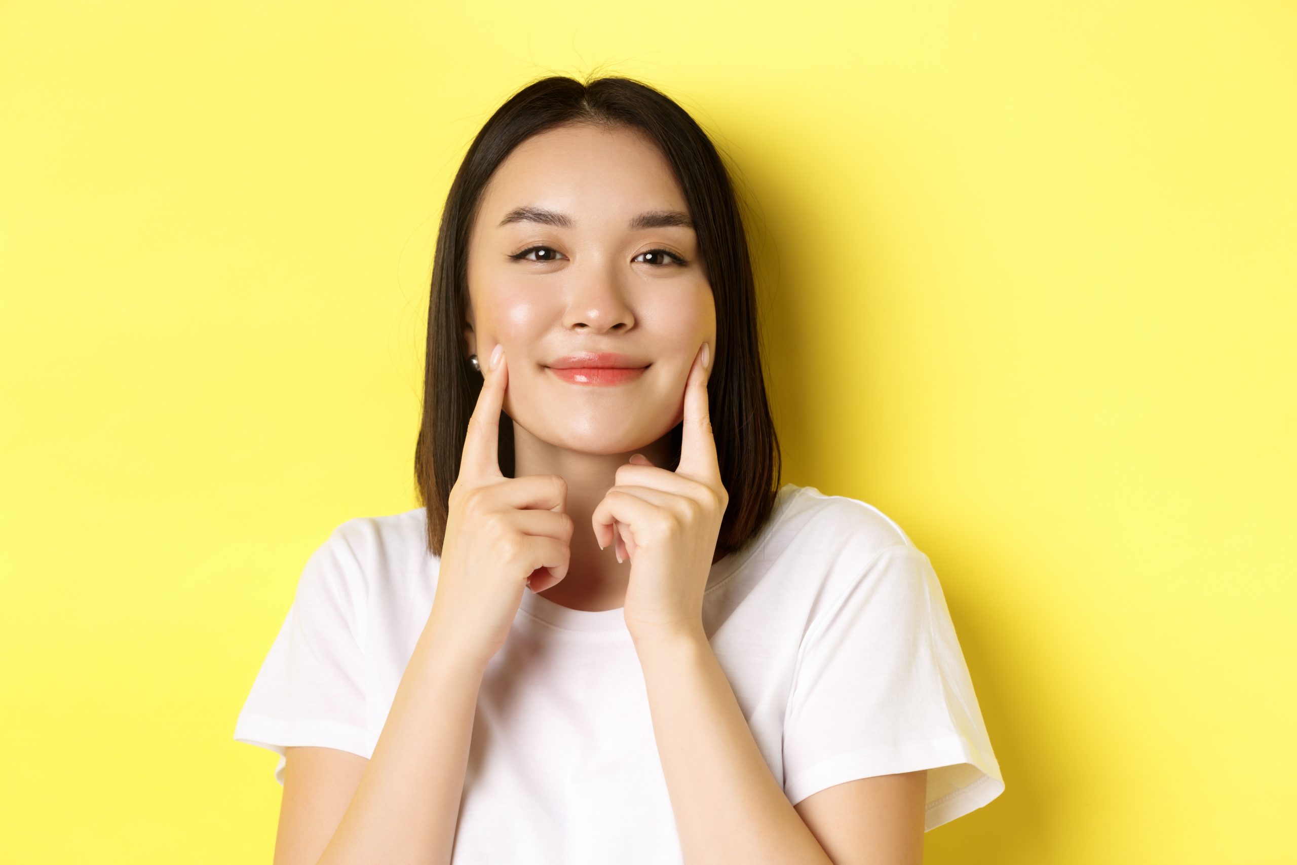 Beauty and skincare. Close up of young asian woman with short dark hair, healthy glowing skin, smiling and touching dimples on cheeks, standing over yellow background